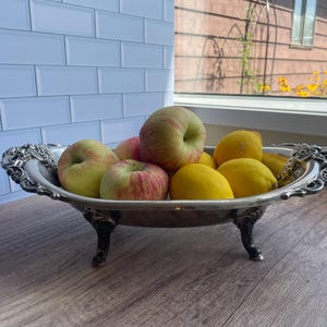 May include: An oval silver-toned metal bowl with ornate, decorative handles and four curved feet. The bowl is filled with a collection of fresh fruit, including several red and yellow apples and bright yellow lemons arranged inside the basin.