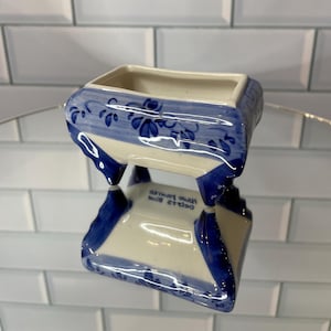 May include: A rectangular ceramic planter with a white base and hand-painted blue floral patterns. The piece features four small feet and the text "HAND PAINTED DELFTS BLUE" printed on the underside, visible through its reflection.