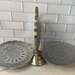 May include: A silver-toned metal serving stand featuring two circular, scalloped-edge trays attached to a central vertical handle. The trays have a decorative cut-out pattern around the perimeter and a textured center, supported by a round base.