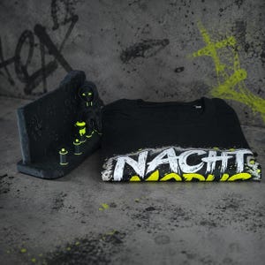 May include: A black t-shirt featuring the white text NACHT and yellow text MODUS with a distressed paint splatter effect. Beside it stands a small black figurine wearing a hooded jacket with neon yellow accents and three miniature spray paint cans.