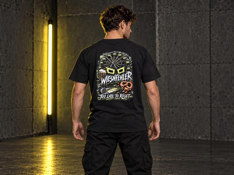 May include: A black short-sleeve t-shirt featuring a graphic on the back with a Ferris wheel, a beer mug, and a pretzel. The text reads WIESNFEHLER and TOO LATE TO RESET in white and yellow lettering against a dark, detailed background illustration.