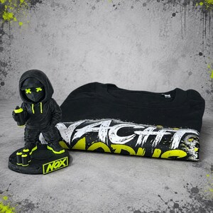 May include: A black figurine wearing a hoodie and mask with neon yellow X-shaped eyes, holding a spray can, standing on a base labeled NOX. Beside it is a black t-shirt featuring white and neon yellow distressed text that reads NACHT MODUS.