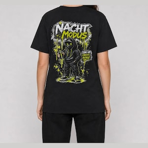 May include: A black t-shirt featuring a graphic on the back of a hooded figure wearing a mask with X-shaped eyes, holding a spray paint can. The text reads NACHT MODUS and STAY UP. CREATE. REPEAT. with yellow and white paint splatter accents.