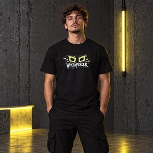 May include: A black short-sleeved t-shirt featuring a graphic print of lime green stylized glasses above the white text WIESNFEHLER. The design includes white paint splatter accents surrounding the glasses and text on the chest area.
