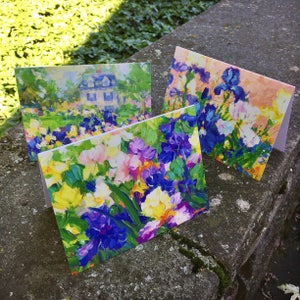 May include: Three greeting cards with colorful floral paintings. The cards feature impressionistic art of irises and a house. The paintings use a palette of blues, purples, yellows, and greens. The cards are displayed on a stone surface.