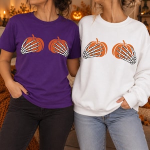 May include: A purple short-sleeved t-shirt and a white long-sleeved sweatshirt, both featuring a graphic print of two orange pumpkins being held by two white skeleton hands. The garments are displayed side-by-side, showcasing the identical design.