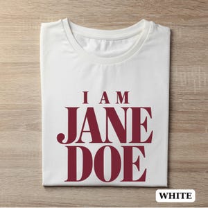 May include: A white short-sleeved t-shirt featuring the text I AM JANE DOE printed in a bold, dark red serif font across the center of the chest. The garment has a classic crew neckline and is presented in a flat, folded orientation.