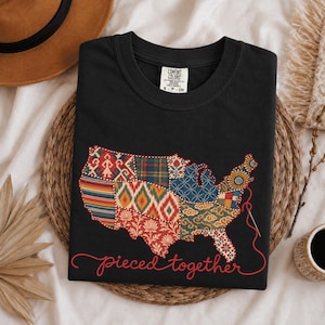 May include: A black t-shirt featuring a graphic of the United States map filled with various colourful patterned fabric swatches. A red thread and needle are attached to the map, with the text "pieced together" written in red cursive script below.
