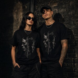 May include: A black short-sleeved t-shirt featuring a graphic print of two faces in profile, separated by a vertical line with a textured, distressed aesthetic. The design is rendered in shades of grey and white, centred on the chest of the garment.
