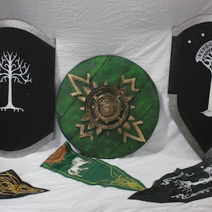May include: Two black heater shields featuring white tree emblems, one with a central star design, and a circular green shield with a metallic gold centre. Three fabric banners are displayed below, featuring white tree and horse motifs in black and green.