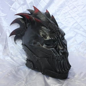 Op de afbeelding: Een zwarte integraalhelm met een schedelachtig ontwerp, diepliggende oogkassen en een gestikt mondgedeelte. De helm heeft een reeks scherpe, vinachtige uitsteeksels aan de boven- en achterkant, geaccentueerd met subtiele rode randen.