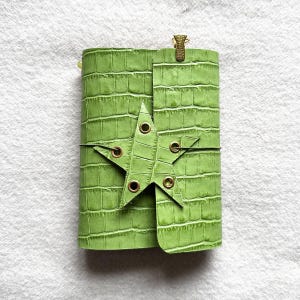 May include: A lime green journal with a textured crocodile skin pattern. The cover features a star-shaped flap closure secured with a thin cord threaded through brass-colored metal eyelets and a small brass-colored metal clip at the top.