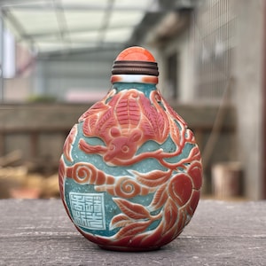 May include: A small, rounded snuff bottle featuring a teal-coloured body with intricate, raised relief carvings in a reddish-orange hue. The design depicts a bat and foliage, with a square seal containing Chinese characters and a bright orange stopper.