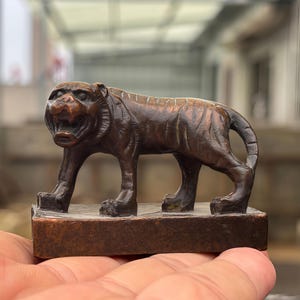 May include: A small, dark brown, metallic tiger figurine standing on a rectangular base. The tiger is depicted in a prowling stance with its mouth open, showing teeth, and its body is covered in engraved stripes. The object has a weathered finish.