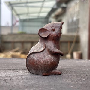 May include: A small, dark brown, textured figurine of a mouse in a seated, upright position. The mouse has rounded ears, a pointed snout, and a smooth, curved tail that wraps along its side. The surface of the figurine has a granular appearance.