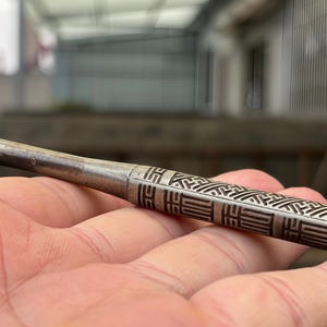 May include: A metallic smoking pipe with a slender, tapered stem and a cylindrical body featuring an intricate, engraved geometric pattern of repeating rectangular and diagonal lines. The surface has a weathered, dark gray finish with visible texture.