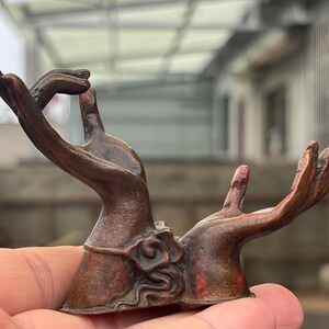 Peut inclure: Une sculpture en métal de couleur bronze représentant deux mains tendues vers le haut. La main gauche est positionnée avec les doigts repliés dans un geste, tandis que la main droite est ouverte et étendue. La base présente un motif en relief.