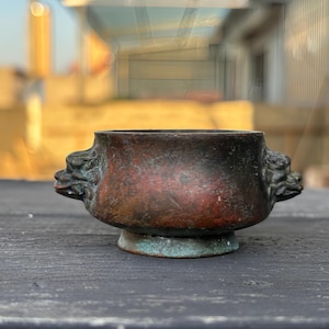 May include: A round, dark bronze-toned metal bowl featuring two sculpted animal head handles on opposite sides. The surface displays a weathered, mottled patina with shades of brown, black, and hints of oxidized green near the circular base.
