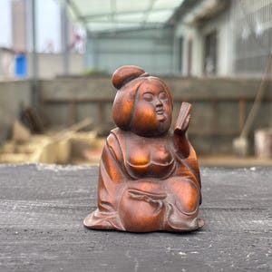 May include: A carved wooden figurine of a seated figure with a rounded face and hair styled in a topknot. The figure wears a robe with draped sleeves, holds a small rectangular object in one hand, and has a smooth, polished brown finish.