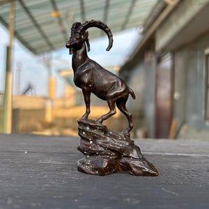 Könnte beinhalten: Eine dunkel bronzefarbene Skulptur eines Steinbocks, der auf einem felsigen Sockel steht. Die Bergziege hat lange, gebogene, geriffelte Hörner und ist im Profil dargestellt. Der Sockel weist kleine weiße Sprenkel auf der Felsstruktur auf.