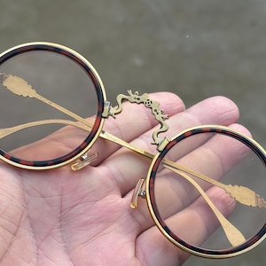 May include: Round spectacles featuring a gold-toned metal frame with a tortoiseshell pattern rim. The bridge is designed with an intricate dragon-shaped metal detail. The thin gold-toned temples have decorative textured accents near the hinges.