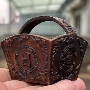 Puede incluir: Un pequeño recipiente de madera tallada en color marrón oscuro con un asa curva. El exterior presenta intrincados relieves de motivos florales y dos medallones circulares, cada uno con un carácter chino, sobre una superficie texturizada.