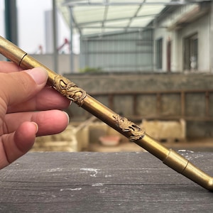May include: A long, slender brass smoking pipe featuring intricate, raised relief carvings of dragons along the stem. The pipe has a smooth, metallic finish with a cylindrical mouthpiece at one end and a wide, circular bowl at the other end.