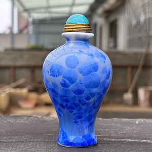 Puede incluir: Una pequeña botella de vidrio con cuerpo redondeado y cuello estrecho, que presenta una base blanca translúcida con patrones cristalinos circulares de color azul vibrante. La botella tiene un tapón de metal dorado con una piedra turquesa lisa.