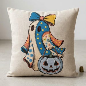 Può includere: Un cuscino quadrato in tessuto beige chiaro con una grafica centrale di un fantasma che indossa un grande fiocco blu e giallo. Il fantasma presenta motivi a pois e floreali blu e tiene in mano un secchiello a forma di zucca jack-o'-lantern.