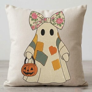 Puede incluir: Un cojín cuadrado de color blanco roto con un gráfico bordado central de un fantasma que lleva un gran lazo floral. El fantasma tiene parches en amarillo mostaza y verde apagado, y sostiene un cubo de calabaza naranja para pedir dulces.