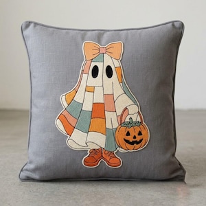 Könnte beinhalten: Ein quadratisches graues Dekokissen mit einem gestickten Aufnäher eines Geistes, der eine pfirsichfarbene Schleife und orangefarbene Turnschuhe trägt. Der Geist hat einen Körper im Patchwork-Stil in gedeckten Orange-, Blau- und Cremetönen und trägt einen Kürbis.