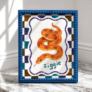 May include: A framed illustration of an orange snake coiled on a white background with a purple scalloped border. The name ziggie is written in blue below the snake. The frame is blue with a beaded texture and a checkered pattern border.