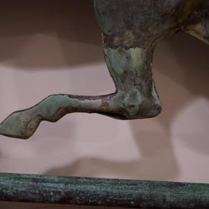 May include: A close-up of a green metal horse sculpture's leg. The horse is in mid-stride, with its leg raised and bent at the knee. The sculpture is made of a textured metal, and the leg is painted in a green color.