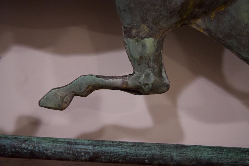 May include: A close-up of a green metal horse sculpture's leg. The horse is in mid-stride, with its leg raised and bent at the knee. The sculpture is made of a textured metal, and the leg is painted in a green color.