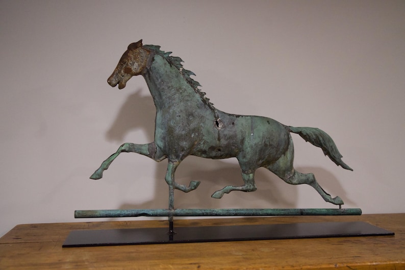 May include: A green metal horse weathervane with a long tail, standing on a black metal base. The horse is in a running pose with its head turned to the side.