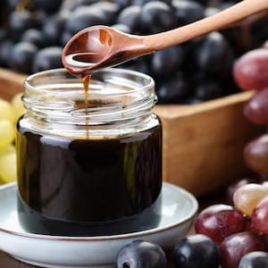 Może przedstawiać: Mały, przezroczysty szklany słoik wypełniony ciemną glazurą balsamiczną, stojący na jasnoniebieskim ceramicznym spodku. Drewniana łyżka znajduje się nad słoikiem, a cienka strużka ciemnego płynu kapie z łyżki do wnętrza naczynia.