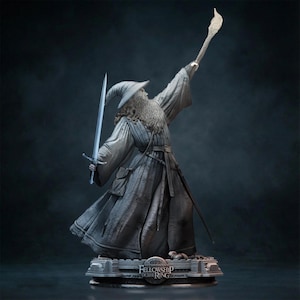 May include: A grey-toned statue of a bearded wizard wearing a pointed hat and long robes, holding a sword in one hand and a glowing staff raised high in the other. The base features the text The Lord of the Rings The Fellowship of the Ring.