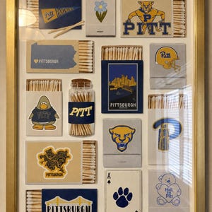 May include: A gold-framed wall art piece featuring a collection of matchboxes and a glass jar filled with matches. The items display various Pittsburgh and Pitt Panthers themed graphics, including a mascot, city skyline, bridges, and a playing card.