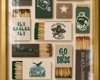 May include: A gold-framed display containing various matchboxes featuring Philadelphia Eagles-themed graphics, including team logos, the text 'FLY EAGLES FLY', 'PHI', 'GO BIRDS', and 'CHAMPIONS PHILADELPHIA', with green-tipped wooden matches inside.