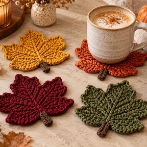 Puede incluir: Cuatro posavasos de ganchillo con forma de hoja en amarillo mostaza, naranja quemado, burdeos oscuro y verde oliva. Cada posavasos presenta un diseño texturizado con nervaduras y un pequeño tallo marrón. Una taza de cerámica descansa sobre uno.