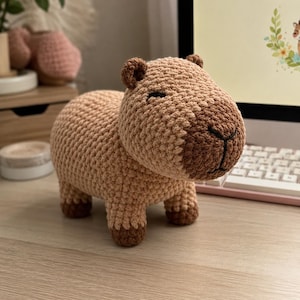 Peut inclure: Une figurine de capybara crochetée en fil beige clair avec un museau et des pattes marron foncé. L'animal en peluche présente de petites oreilles arrondies, des yeux fermés brodés et un nez cousu, debout sur quatre pattes courtes.