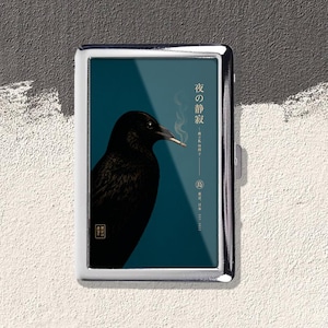 May include: A rectangular metal cigarette case featuring a dark teal background with a black crow holding a lit cigarette in its beak. The design includes white Japanese text, a thin vertical line, and a small circular emblem with a bird character.