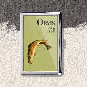 May include: A rectangular metal case with a polished silver frame featuring a light green insert. The insert displays a vintage illustration of a fish on a fishing line with the black text Orvis spring 1969 printed in the upper portion.