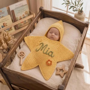 May include: A yellow crocheted star-shaped baby swaddle blanket with a hood, featuring the name Mia embroidered in green cursive script and a small orange flower appliqué. The swaddle is laid flat, surrounding a sleeping infant in a wooden crib.