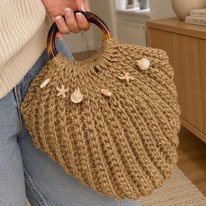 May include: A tan crocheted handbag shaped like a seashell, featuring a round tortoiseshell-patterned handle. The exterior is decorated with several small, light-colored seashells and starfish attached to the textured, ribbed knit surface.