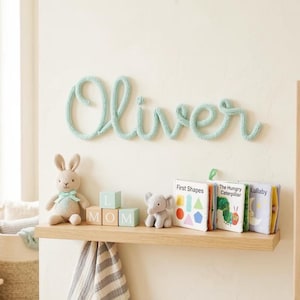 Pode incluir: Um letreiro de nome feito de fio tricotado em verde menta claro que soletra Oliver, montado numa parede acima de uma prateleira de madeira. A prateleira contém um coelho de peluche bege, três blocos de madeira claros que soletram MOM, um pequeno elefante de peluche cinzento e três pequenos livros.