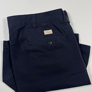 May include: A pair of navy blue chino shorts featuring a waistband with belt loops, a single rear welt pocket with a button closure, and a rectangular beige fabric label stitched above the pocket that reads Polo U.S.A. Ralph Lauren.