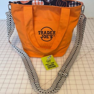 May include: An orange canvas tote bag with the black Trader Joe's logo printed on the front. The bag features a black and white herringbone patterned adjustable shoulder strap and a black interior lining with a white and orange pumpkin print.