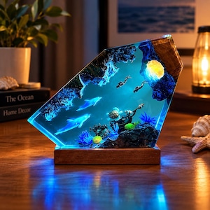 Pode incluir: Uma lâmpada de resina trapezoidal com uma cena subaquática contendo dois golfinhos, uma tartaruga marinha, dois mergulhadores e um navio naufragado. A peça é iluminada com luz azul e assenta sobre uma base retangular de madeira.