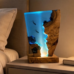 Peut inclure: Une lampe de table rectangulaire avec une base en bois naturel et un corps vertical composé de bois et de résine bleue translucide. La section en résine contient deux petites silhouettes de plongeurs et imite une scène sous-marine.
